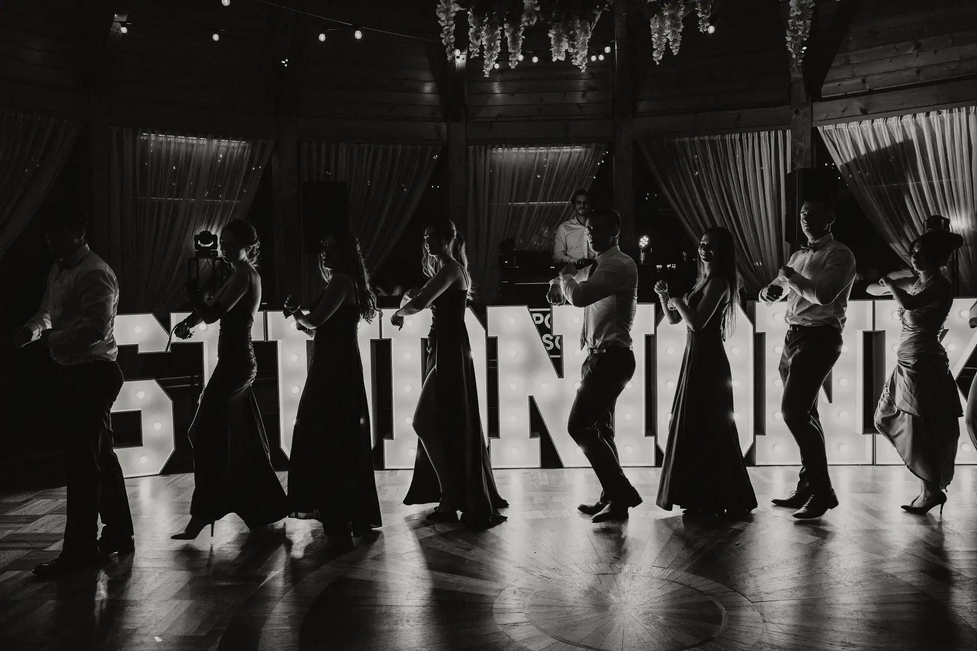 Sylwetki tańczących par przed podświetlonym napisem, w czerni i bieli